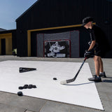 En pojke på Extreme Hockey Flooring Tiles tränar med puckar, framför en målvaktsskydd och en hockeymålvaktsskott. Perfekt plastis hockeygolv.