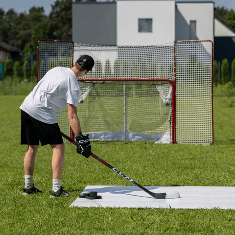 En spelare övar skott med en hockeyklubba framför ett hockeymål i gräset, med puckar framför sig. Perfekt för träning hemma.