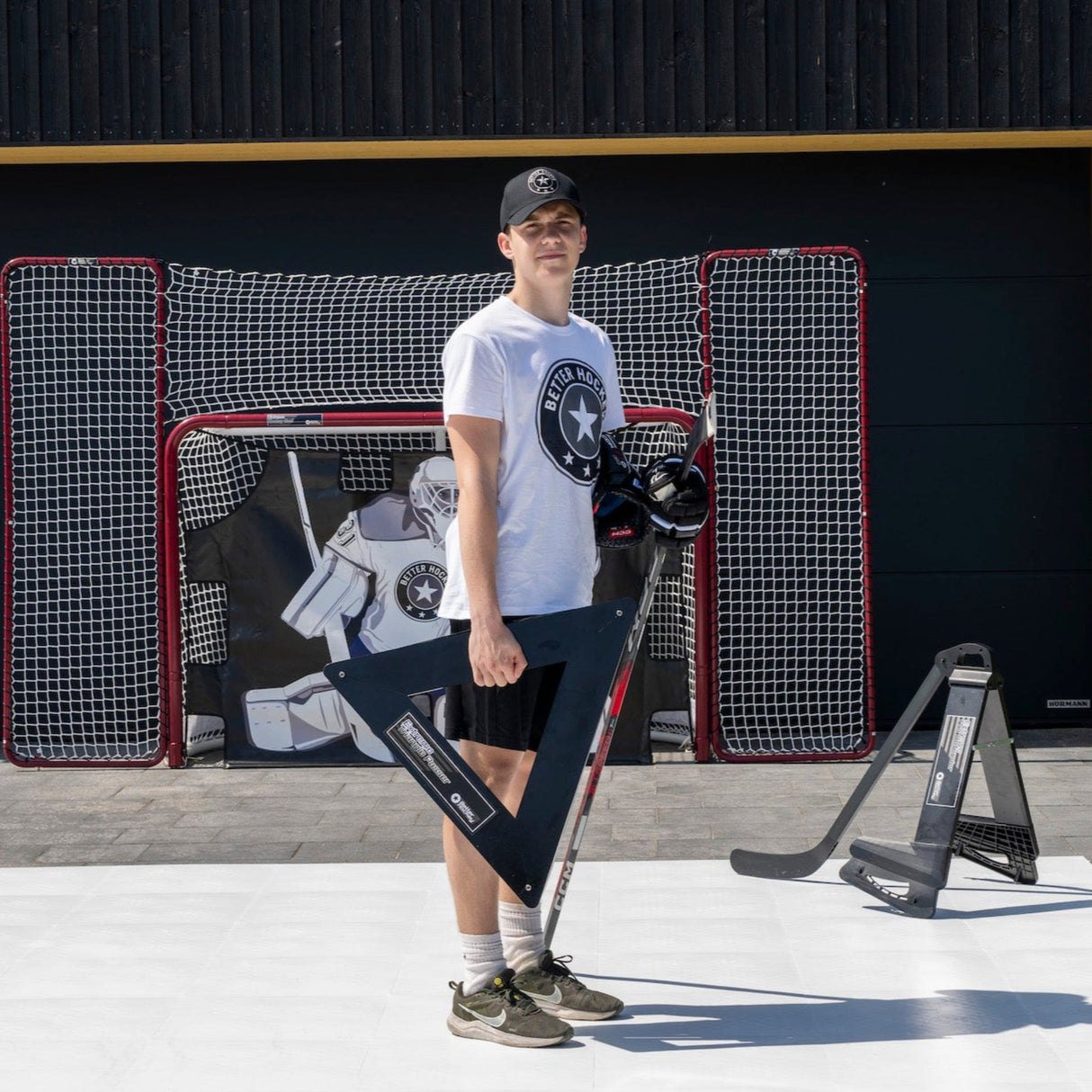 En man står med en extrem hockeypassare i hand, klädd i t-shirt och keps, med hockeymål i bakgrunden.