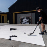 Ung kille övar hockeypassning med puckar på en specialdesignad yta, med en passer och målvaktsmål i bakgrunden.